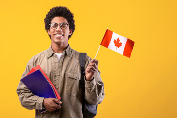 Education, school, college and university. Happy young african american guy in glasses holding notepads and small Canada flag and ready to study, isolated on orange background, free space, studio shot