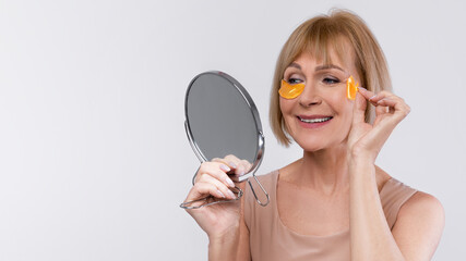An elderly woman applies eye patches while smiling at her reflection in a mirror. She indulges in...