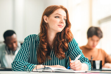 Portrait of smiling beautiful redhead female university student holding pen writing in notebook...