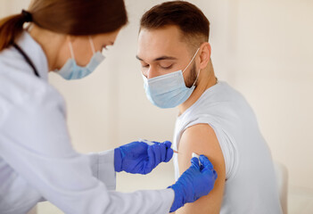 Female doctor making injection of coronavirus vaccine to young patient in medical mask at health...