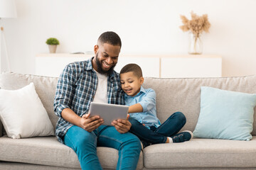 Young Black Father And Little Son Using Tablet Browsing Internet Together And Watching Movie Online Sitting On Sofa At Home On Weekend. Family, Gadgets. Daddy And Child Using Computer Concept