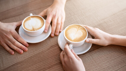 Cropped view of young couple having coffee with heart shaped foam at table in cafe. Unrecognizable millennial guy with his girlfriend having romantic date at restaurant, enjoying tasty hot beverage