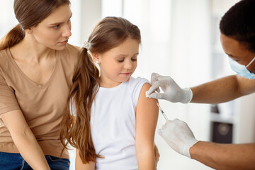 Doctor doing injection of covid-19 vaccine to little patient at clinic. Cute girl with her mom on...