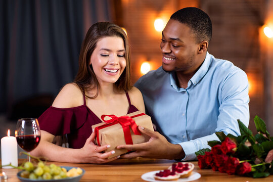 Valentine's Day Celebration. Happy Romantic Interracial Couple Dining In Restaurant, Exchanging Gifts, Loving Black Man Giving Wrapped Present Box To His White Girlfriend, Greeting With Anniversary