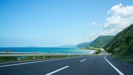 Scenic coastal highway curving along a lush green mountainside beside a vibrant blue ocean under a clear sky
