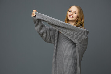 Young girl smiling in oversized gray sweater, showcasing cozy fashion against a muted background,...