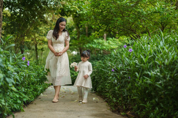 happy mother and toddler girl in white dress walking in flower garden
