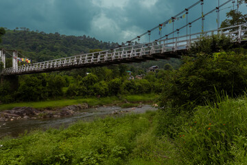 An elegant suspension bridge crosses a gently flowing river in himachal pradesh, india, surrounded by dense greenery and distant mountains beneath a dramatic cloudy sky.