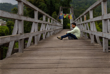 Man sitting on wooden bridge in lush green forest. A thoughtful man sits quietly on a wooden bridge amidst lush vegetation in Himachal Pradesh, India.