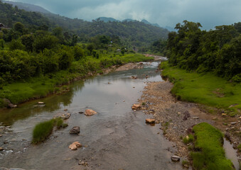 A calm river flows gently between lush, green forested mountains under a canopy of overcast skies...