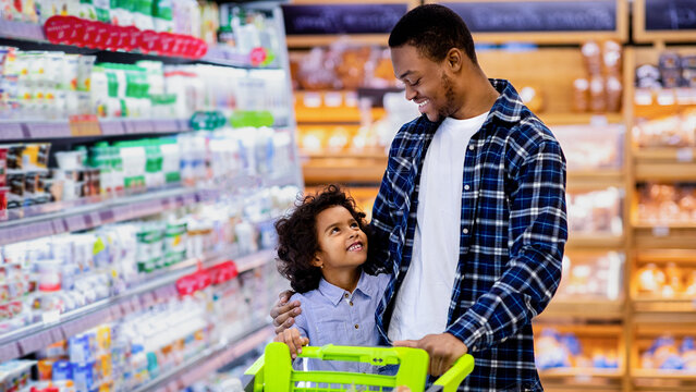 Happy black dad and his pretty little daughter with shopping trolley having great time buying groceries at big mall. African American parent with kid purchasing foodstaff at hypermarket