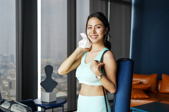 Smiling Asian girl in sportswear with yoga mat using towel to wipe sweat after yoga practice exercise inside residence. Young sporty woman taking a break after home gym indoor, Workout sport concept