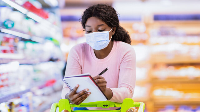 African American woman in face mask doing shopping at supermarket, adding products to grocery list, writing in notebook. Black female customer purchasing goods at big mall