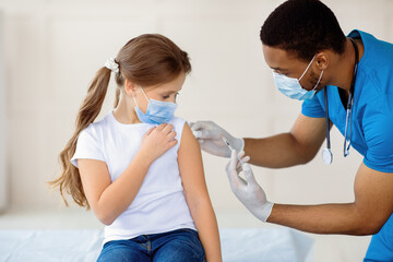 Young black doctor making covid-19 vaccine injection to little girl in face mask at hospital. Child getting coronavirus immunization at clinic. Protection against global virus concept