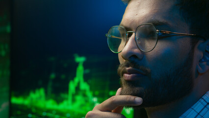 Fototapeta na wymiar Indian Arabian man in glasses thinking contemplate ponder solving coder trader IT specialist male ethnic guy businessman business programmer analysing trading chart graph crypto currency neon indoor