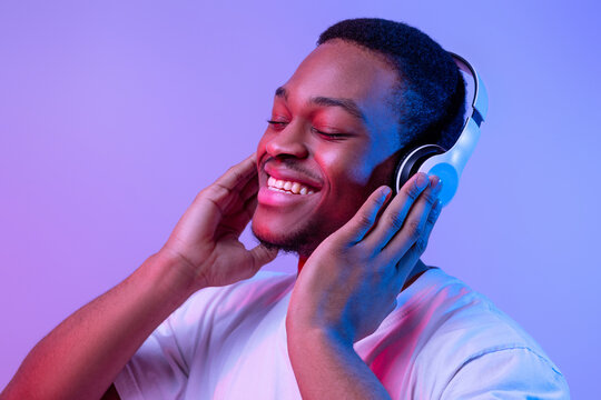 Music Lover. Handsome Black Man Listening Favorite Songs In Wireless Headphones, Enjoying New Playlist, Relaxed African American Guy Standing With Closed Eyes Under Neon Lighting Ove Purple Background