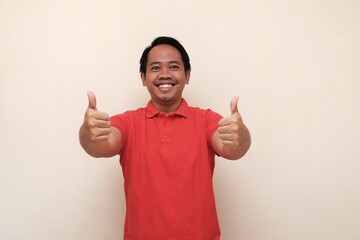 Cheerful Indonesian man in red shirt smiling confidently and giving two thumbs up, expressing approval, support, and celebration.