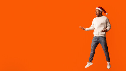 Shocking Christmas Offer. Dazed Black Guy In Santa Hat Looking At Copy Space With Excitement, Astonished African Guy Jumping In Air On Orange Studio Background, Emotionally Reacting To Seasonal Sales