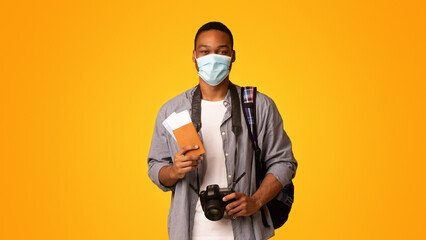 Pandemic Tourism. Happy young african american tourist guy in medical face mask holding passport with tickets and photo camera, ready for vacation, standing isolated over yellow background, copy space