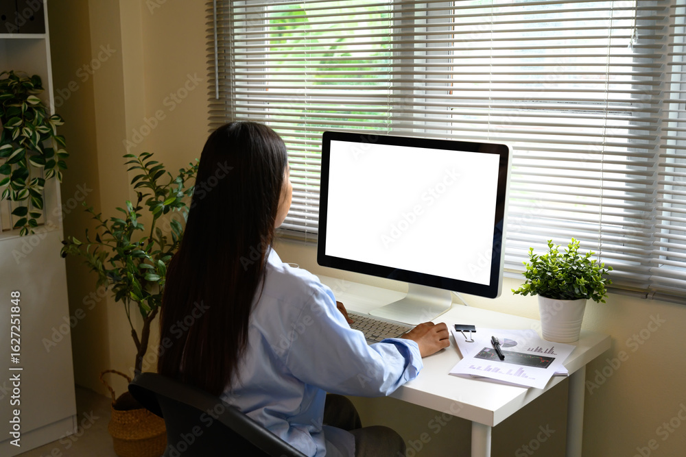 Poster rear view of woman using computer at a desk with documents in a modern workspace