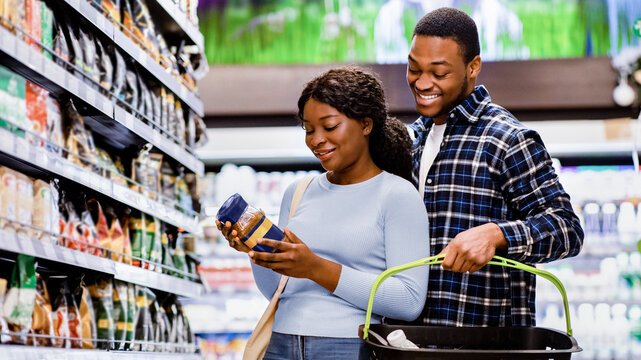 Young African American couple buying groceries, choosing good coffee at supermarket. Cheerful black family purchasing food at store or huge mall, holding basket full of products