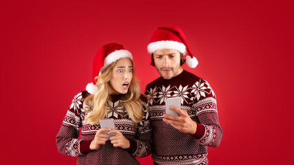 Online Christmas shopping. Shocked millennial couple staring into smartphones on red background