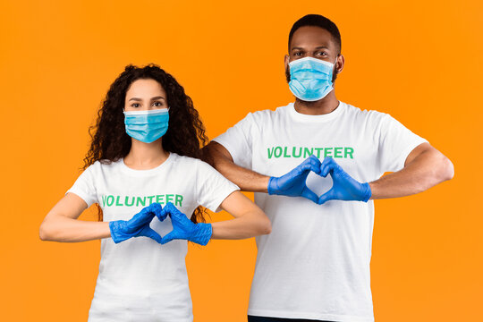 Two Multiethnic Volunteers Gesturing Heart Shape Wearing Protective Face Masks And Medical Rubber Gloves For Coronavirus Protection Posing On Yellow Studio Background. Kindness And Charity Concept - Powered by Adobe