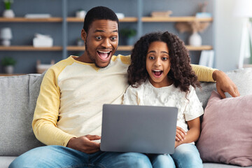 Shocked black dad and daughter using computer while sitting together on couch in living room....