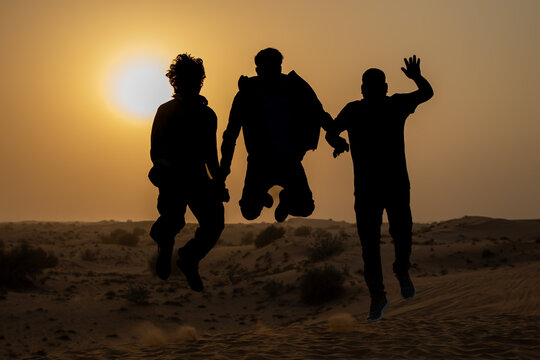 Three people joyfully jump atop a desert dune in Dubai, UAE, during the evening, expressing happiness and excitement. The scene creates stunning silhouettes against the vibrant sunset sky.