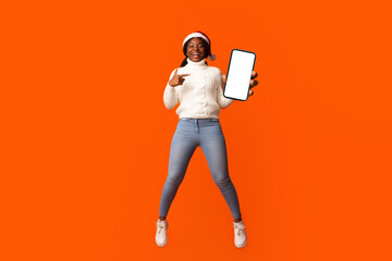 Christmas Offer. Smiling African Lady In Santa Hat Pointing At Smartphone With Black Screen In Her...