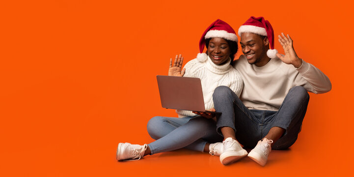 Young black spouses in santa hats making video call on laptop computer, sitting on floor on orange studio background, waving hands at camera, enjoying online communication during christmas holidays