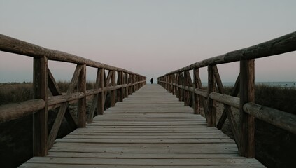 Wooden bridge leading to the horizon. A lone figure walks towards the distant, muted sunset