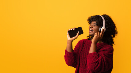 Joyful curly african american woman with smartphone and wireless headset singing on yellow studio...