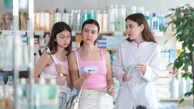 Attentive mother and daughter buyers choosing medical ointment and professional female parmacist helping in small pharmacy 