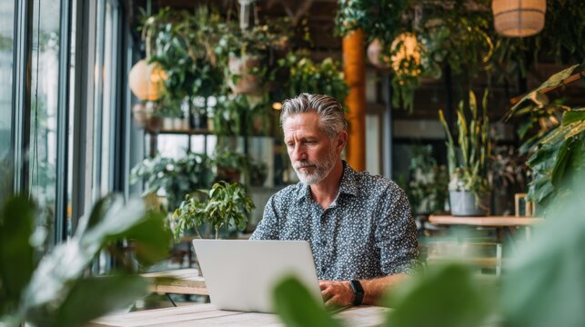 A man with gray hair is focused on his laptop, seated at a table surrounded by lush greenery in a bright cafe during daylight.
