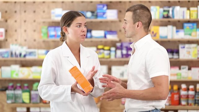 Positive woman apothecary speaking to male customer holding medicinal preparation in box in drugstore