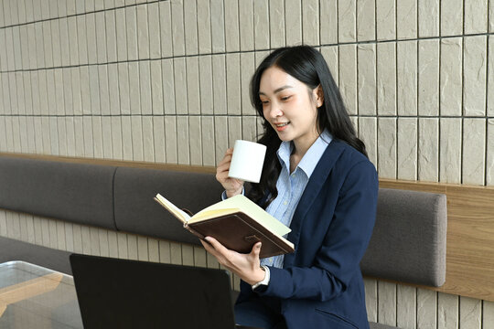 A woman is sitting on a bench with a book and a cup of coffee. She is wearing a blue jacket and she is reading the book while sipping her coffee.