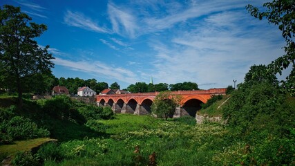 The historic red brick bridge in Kuldīga, Latvia, spanning across the lush green banks of the Venta River, with charming old buildings and a church spire in the background under a bright blue sky.
