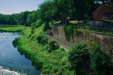 The Venta River in Kuldīga, Latvia, with lush greenery, historic stone walls, and traditional houses with red roofs in the background.