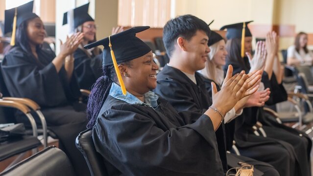 Young people in graduation gowns listen to a speech and applaud. 