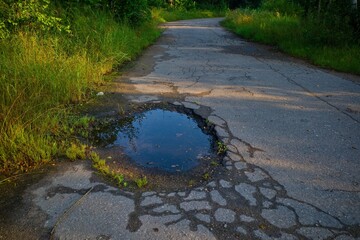A pothole filled with water on a cracked asphalt road surrounded by grass and trees in the evening sunlight.