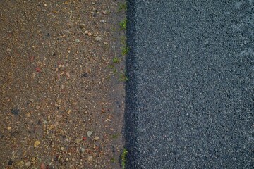 Close-up of the boundary between gravel and fresh asphalt with small green plants growing along the edge.