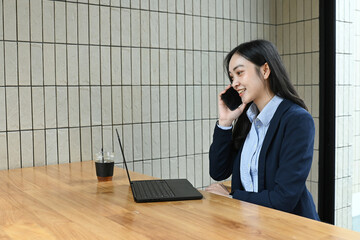 A woman in a business suit is talking on her cell phone. She is sitting at a table with a laptop...