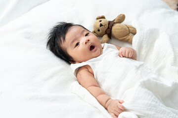 Cute Asian newborn baby wrapped in a white blanket, lying beside a soft brown teddy bear and...
