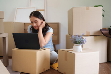 Fototapeta premium Asian young adult woman working laptop computer planning new home relocation organizing apartment surrounded cardboard moving boxes