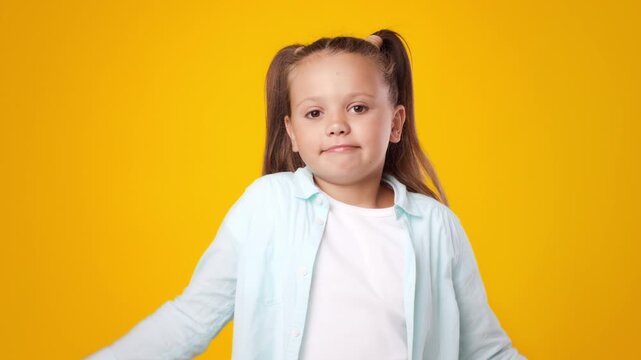 A cheerful child stands in front of a vivid yellow backdrop, shrugging her shoulders in a carefree manner, capturing the essence of happiness and curiosity during a fun moment in childhood.