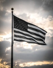 A grayscale photo of an American flag waving in a cloudy sky, backlit by the sun