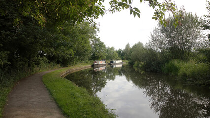 a view down a canal towpath with two narrow boats moored in the distance with reflections in the ...