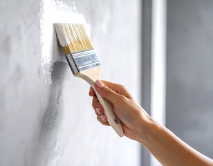 A hand holds a paintbrush applying white paint to a textured grey wall