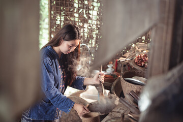 Seen through a frame, a young woman focuses intently on stirring a steaming pot in her rustic kitchen. This intimate shot highlights the dedication required for traditional cooking.